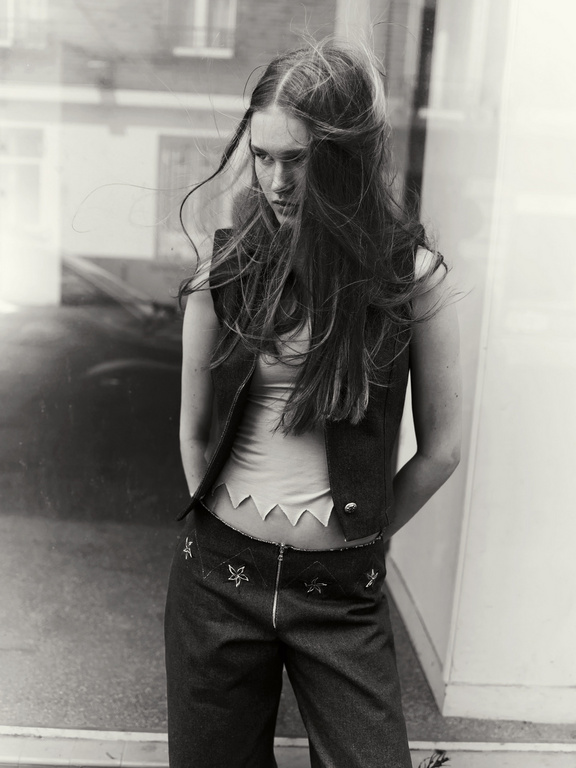 Monochrome portrait of a young woman with long tousled hair wearing a sleeveless vest and trousers outdoors.