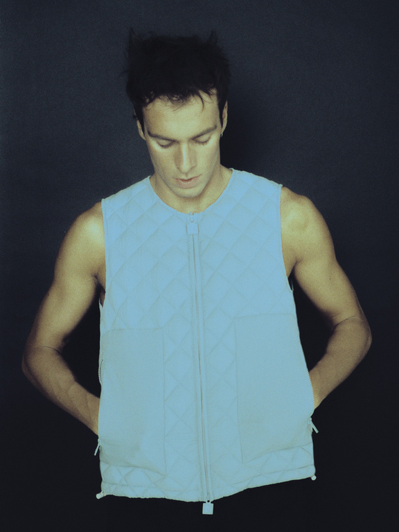 An adult male wears a light blue sleeveless quilted vest with a zipper, hands in pockets, against a dark gray background in a studio shot.