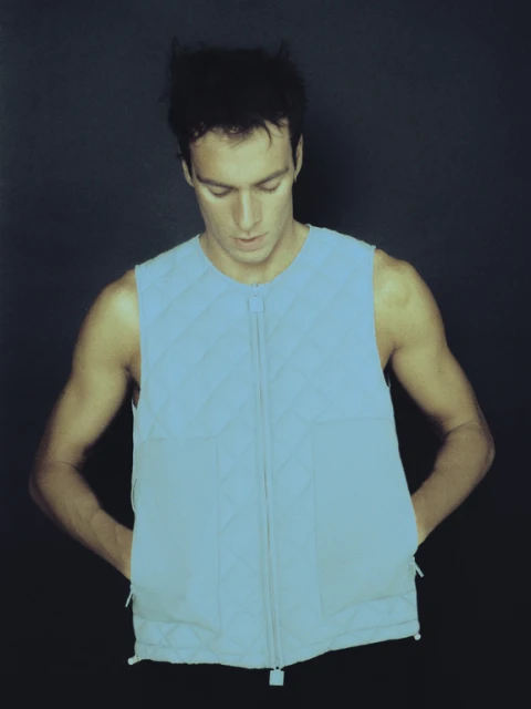 An adult male wears a light blue sleeveless quilted vest with a zipper, hands in pockets, against a dark gray background in a studio shot.