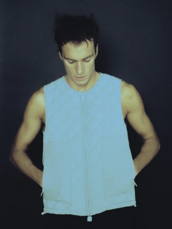 An adult male wears a light blue sleeveless quilted vest with a zipper, hands in pockets, against a dark gray background in a studio shot.