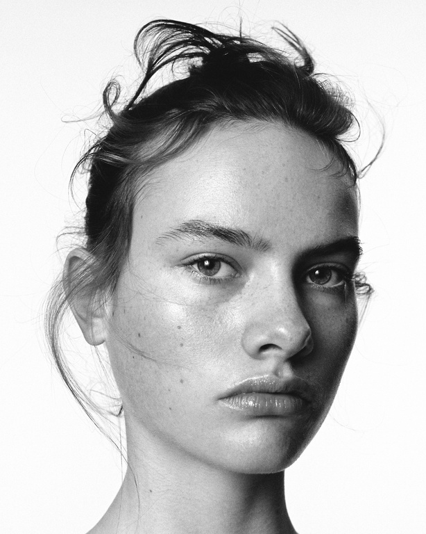 Striking black and white portrait of a young woman with a neutral expression, captured in a photo studio with soft, diffused lighting.