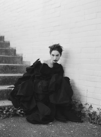 Model with dark curly hair wearing a voluminous black dress seated outdoors on concrete steps next to a brick wall.
