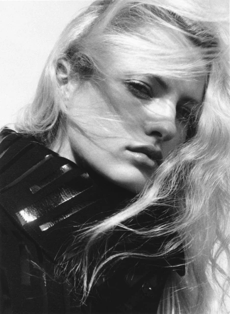Monochrome portrait of a woman with long blonde hair, wearing a black leather collar with a white background, shot at eye-level.