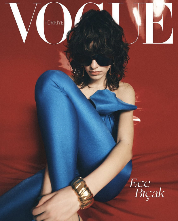 Model wearing blue satin trousers and oversized sunglasses seated against a vibrant red backdrop