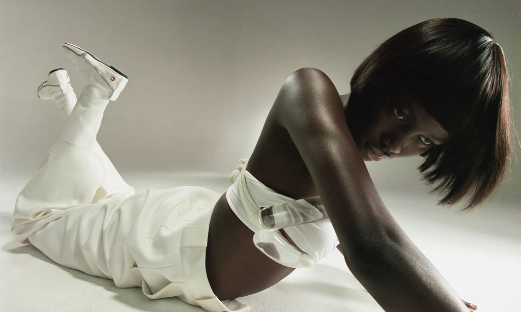 Portrait of a woman in white pants and crop top laying on her side in a studio with white seamless backdrop and wearing white shoes.