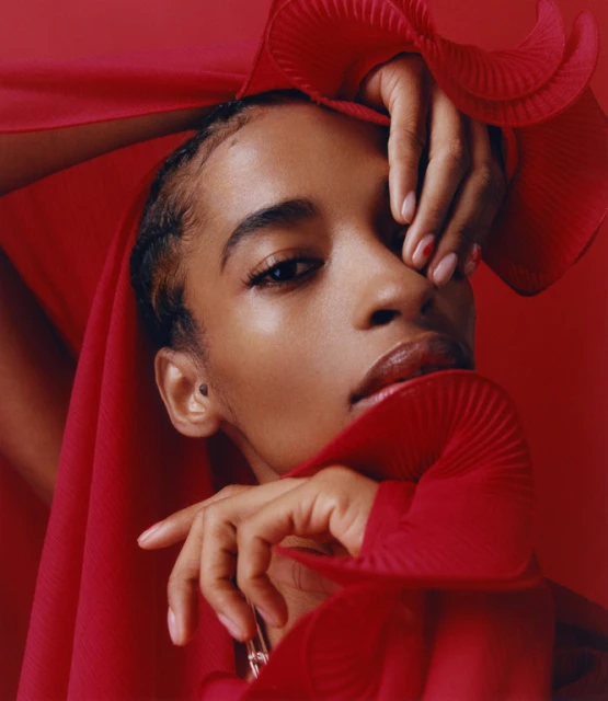 Portrait of a woman with light skin, wearing a red ruffled garment, with her hands near her face, against a solid red background.