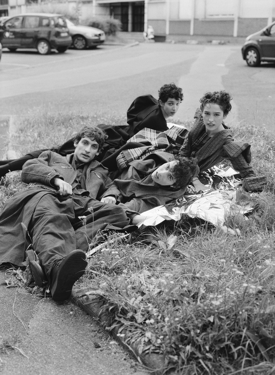 Four models lying in grass near a parking lot with cars, styled by Jacob Kajrup in monochrome editorial photography