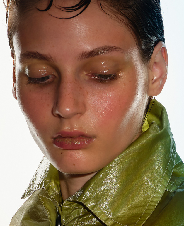 Close-up portrait of a young adult woman with short, dark wet hair, wearing a shiny green jacket, against a bright white background.
