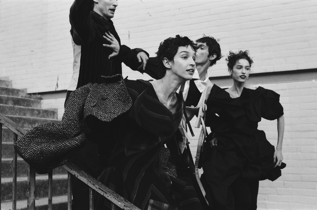 Black-and-white fashion shot of four models in expressive poses and unique clothing on steps against a textured wall backdrop.