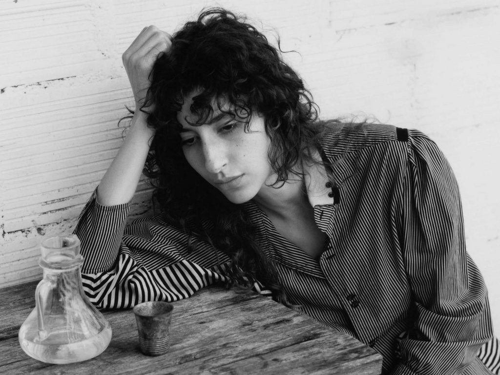 Black and white studio shot of a woman with curly hair leaning on a wooden table with a glass bottle and metal cup in view.