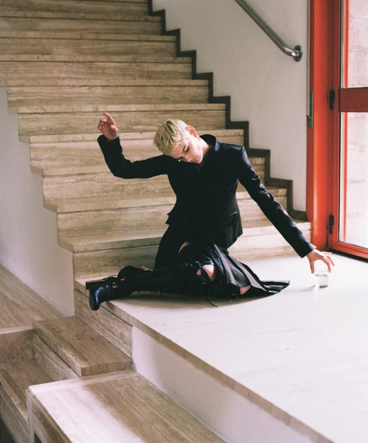 A young adult woman with short blonde hair is kneeling and posing indoors wearing a black jacket and skirt with black boots.