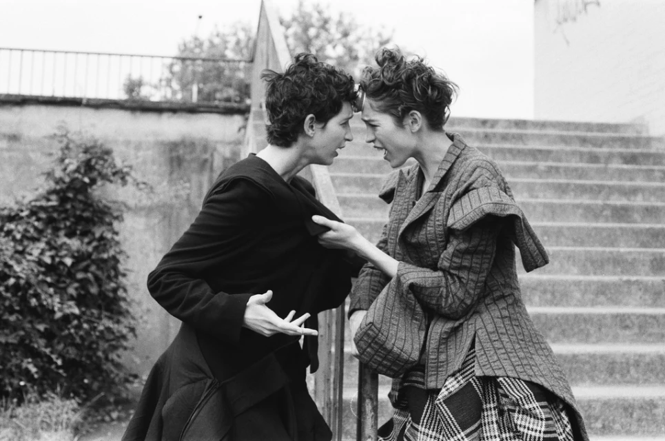 Two adult women facing each other on an outdoor staircase, dressed in layered vintage-style clothing, captured in black and white.