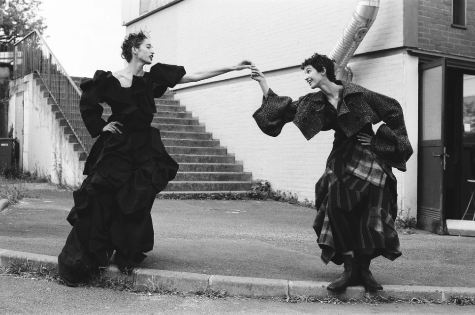 Two women in oversized black clothing pose on a street next to a white building with stairs in a monochrome fashion editorial.