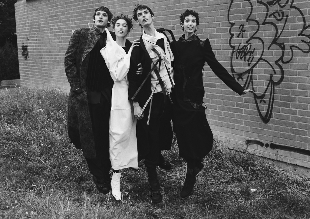 Four fashion models pose in black and white, with one woman in white, against a brick wall with graffiti in an outdoor, edgy scene.