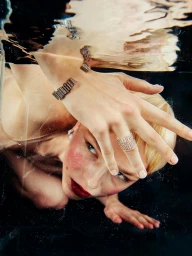 Close-up underwater portrait of a woman with blonde hair wearing jeweled rings and a cuff bracelet near the surface.