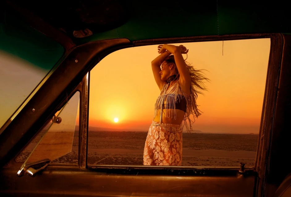 Woman standing in car window at sunset in desert. The vehicle interior frames the outdoor scene in warm, golden hour lighting.