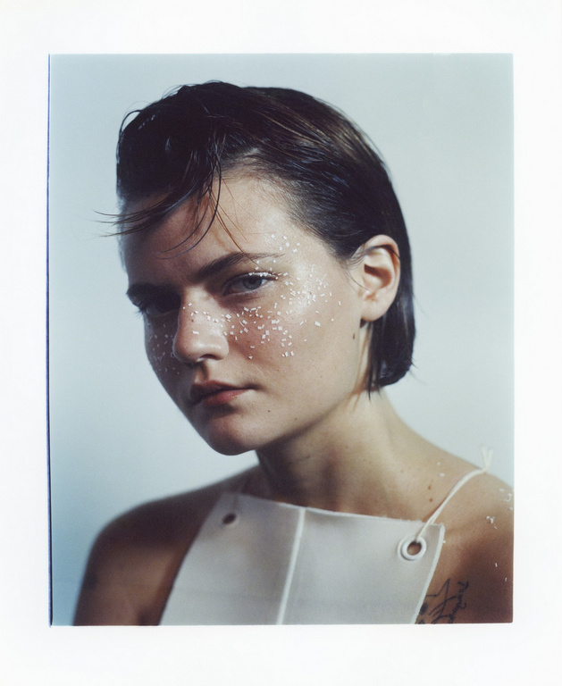 Portrait of a young woman with short dark hair, glitter on her face, and wearing a structured white top with grommets.