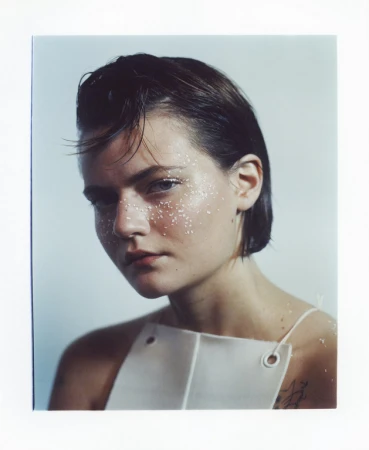 Portrait of a young woman with short dark hair, glitter on her face, and wearing a structured white top with grommets.