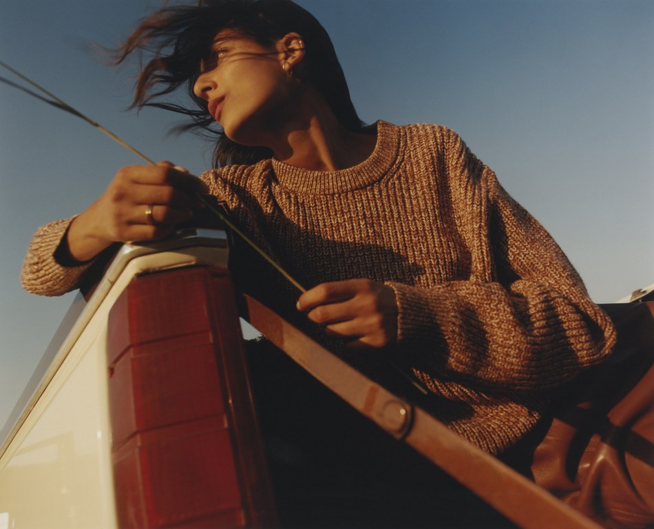 Woman in sweater looks out from a truck bed. She holds grass in her hand as the wind blows through her hair against a blue sky.