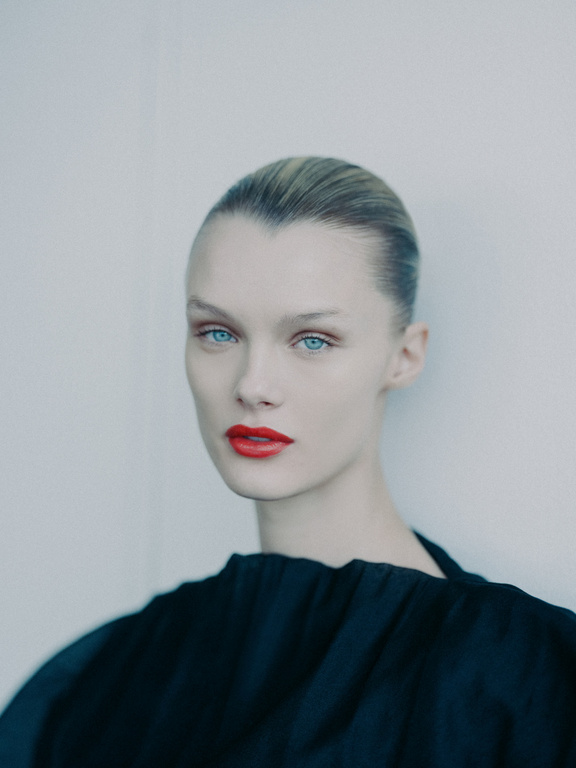 Close-up portrait of a young woman with fair skin, blue eyes, and red lipstick, wearing a black top against a neutral background.