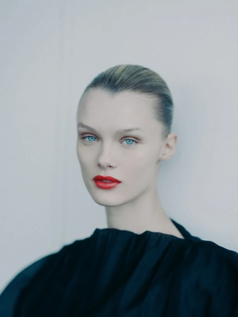 Close-up portrait of a young woman with fair skin, blue eyes, and red lipstick, wearing a black top against a neutral background.