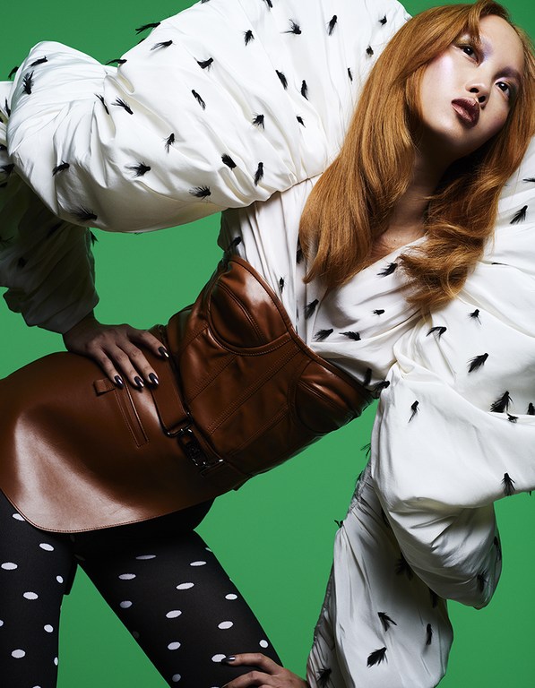 Adult female fashion model poses in a white blouse with feather accents, a brown corset, and polka dot pants against a green backdrop.