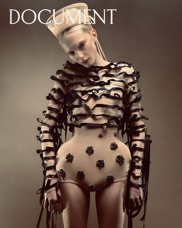 Fashion editorial shot of a woman with pale skin wearing a beige bodysuit with black ribbon detailing on a brown studio background.
