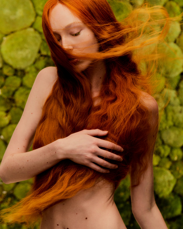 Red-haired woman with eyes closed and hand on her chest poses against a green mossy background in an eye-level medium shot.