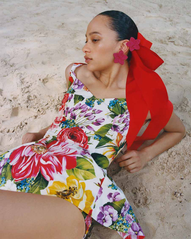 A woman with flower accessories lies on the beach in a floral print bathing suit with a red bow detail, enjoying a day at the shore.