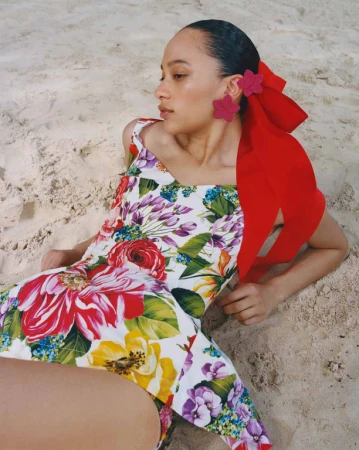 A woman with flower accessories lies on the beach in a floral print bathing suit with a red bow detail, enjoying a day at the shore.