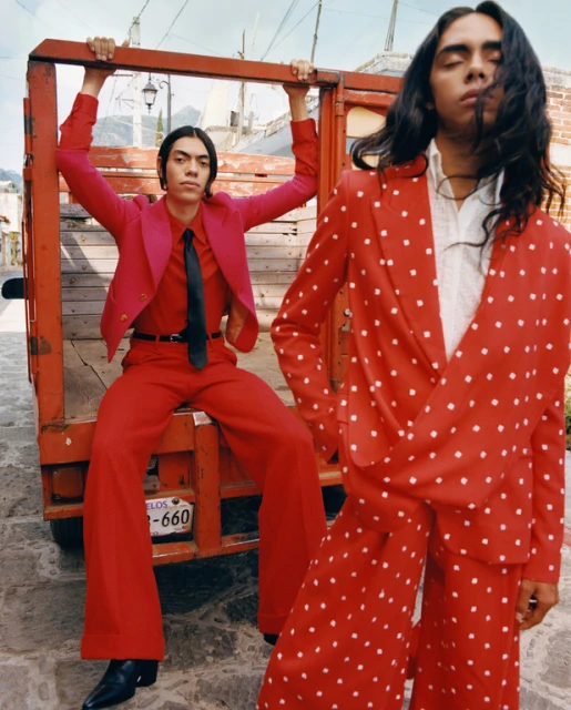 Two fashion models pose on a truck bed in red and pink suits. The background shows an outdoor urban setting with traditional architecture.