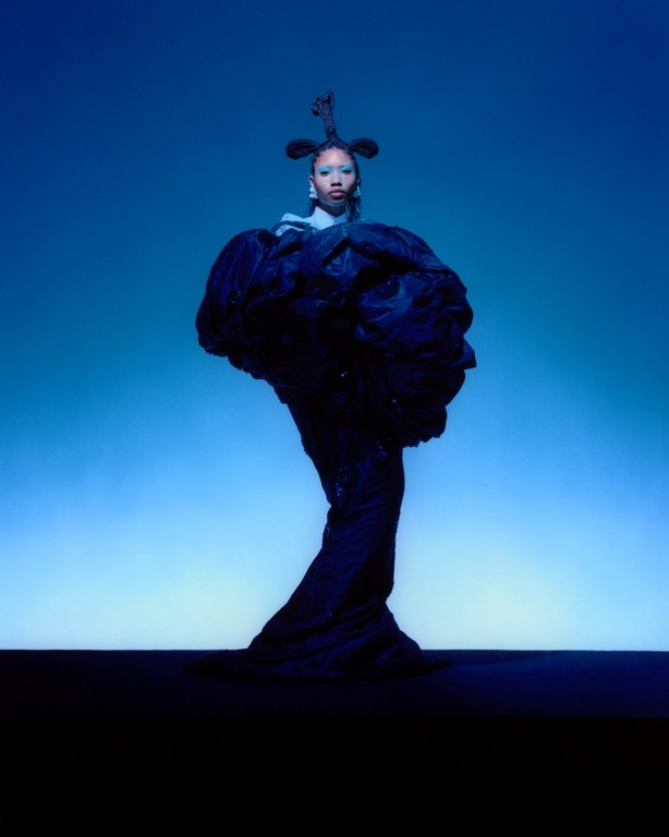 A Black woman stands in a sculptural gown, hair styled high, against a blue gradient background in a fashion photography studio shot.