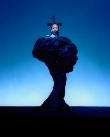 A Black woman stands in a sculptural gown, hair styled high, against a blue gradient background in a fashion photography studio shot.