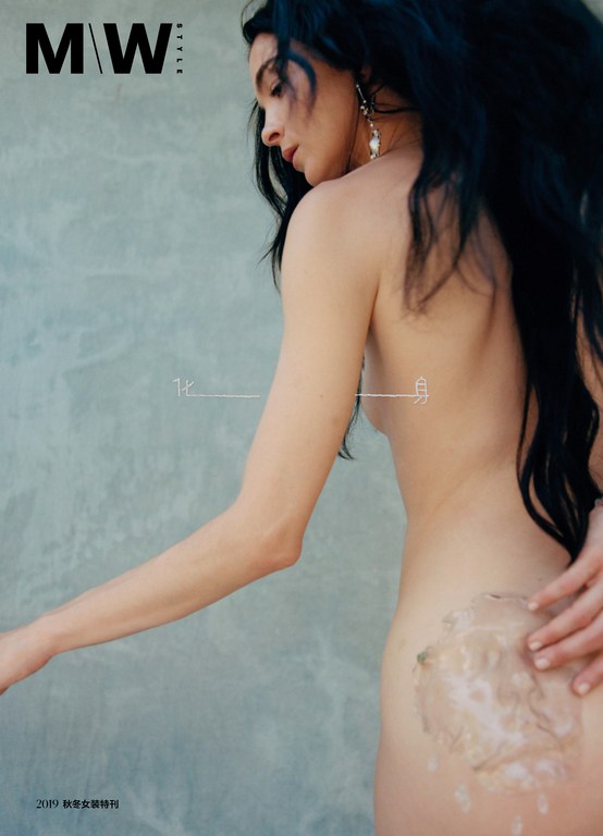 Topless female model poses in studio with long dark hair, pearl earrings, tan skin, hands touching back, in fashion magazine.
