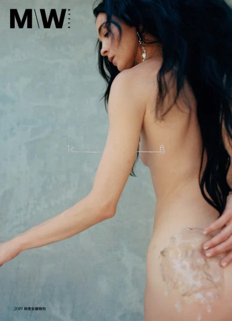 Topless female model poses in studio with long dark hair, pearl earrings, tan skin, hands touching back, in fashion magazine.
