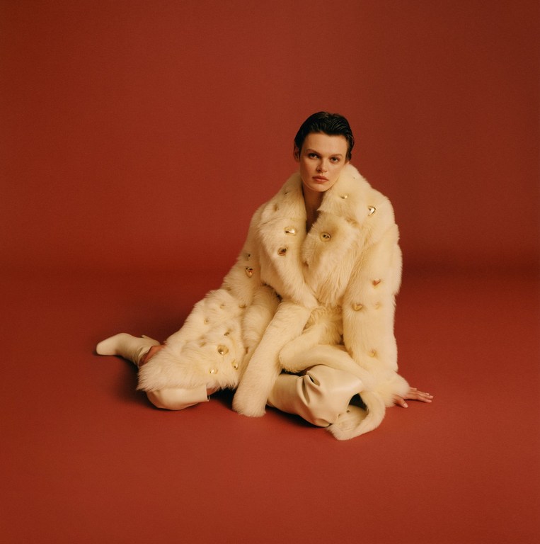 Model seated on a warm terracotta background wearing a plush cream faux fur coat and matching boots.