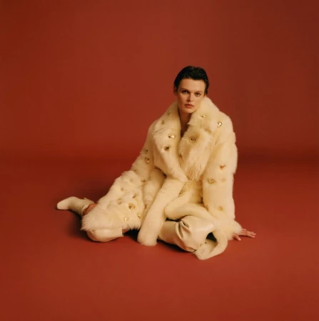 Model seated on a warm terracotta background wearing a plush cream faux fur coat and matching boots.