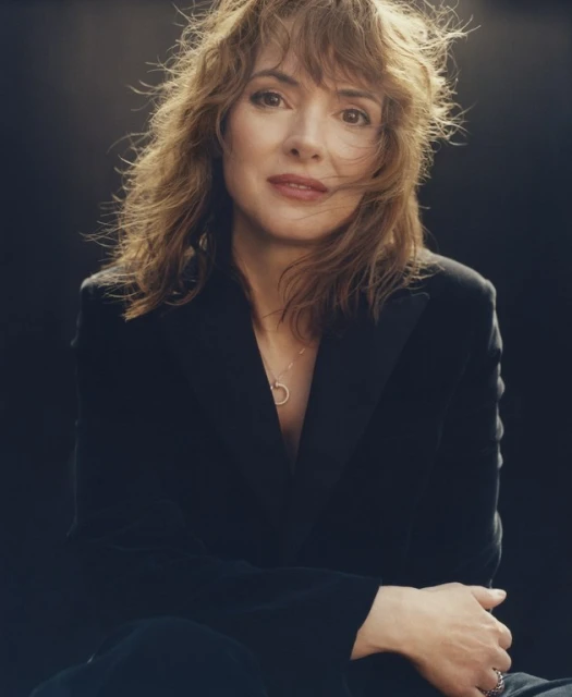 Close-up portrait of an adult woman with shoulder-length wavy hair, wearing a black blazer and necklace against a dark background.
