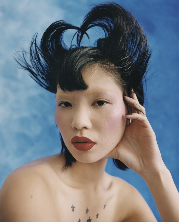 A dramatic beauty shot of an Asian woman with an avant-garde hairstyle, pale eyebrows, and star tattoos, set against a cloudy blue background.