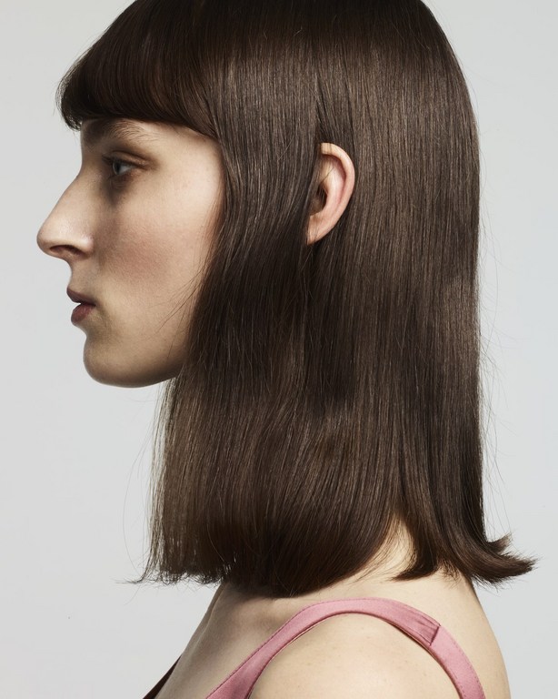 Profile shot of a stylish adult female with sleek, shoulder-length dark brown hair and a pink top against a neutral studio backdrop.