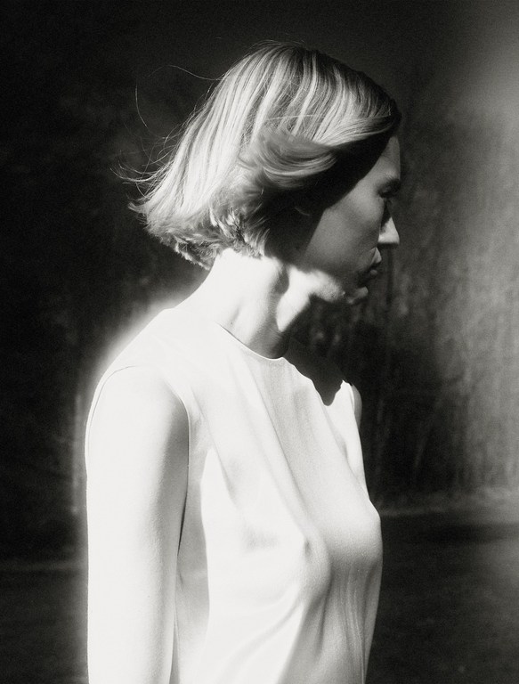 Monochrome portrait of a fair-skinned adult woman with short hair and a white top against a blurred, dark background.