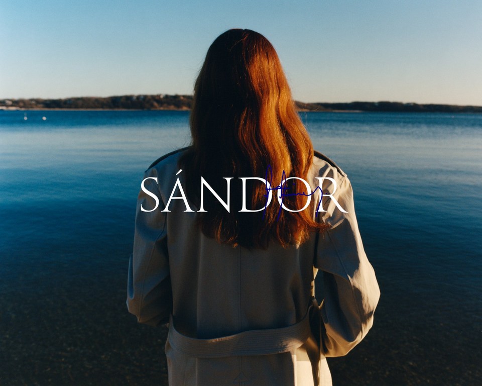 A woman with long red hair stands looking out at a calm body of water on a sunny day. She wears a trench coat.