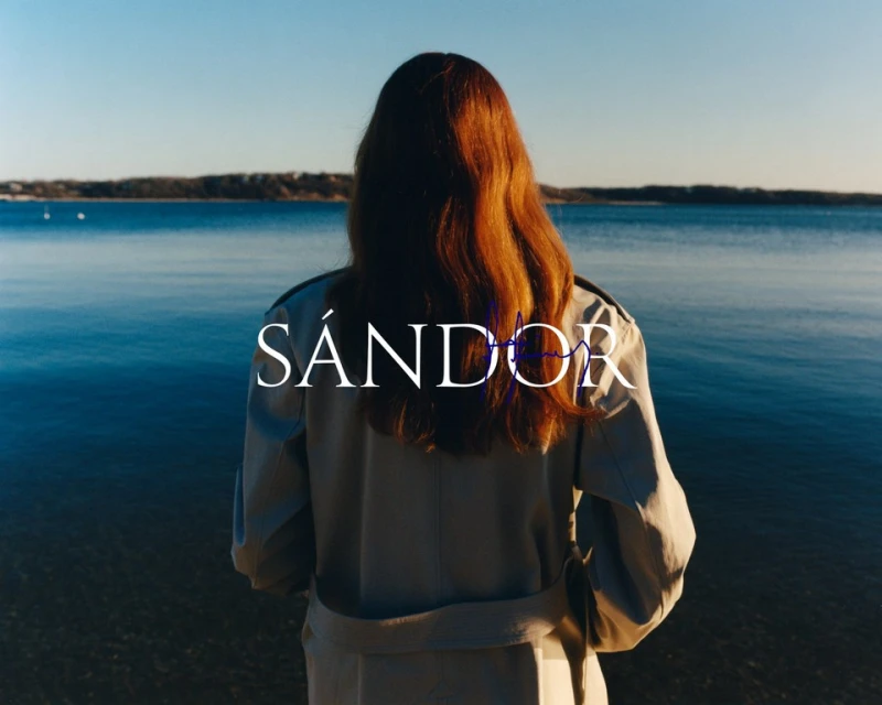 A woman with long red hair stands looking out at a calm body of water on a sunny day. She wears a trench coat.