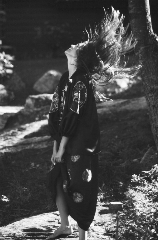 Monochrome portrait of a woman with long hair in motion, wearing an ornate dark dress, posing in a grassy outdoor environment with trees.