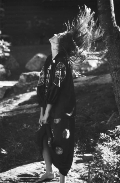 Monochrome portrait of a woman with long hair in motion, wearing an ornate dark dress, posing in a grassy outdoor environment with trees.