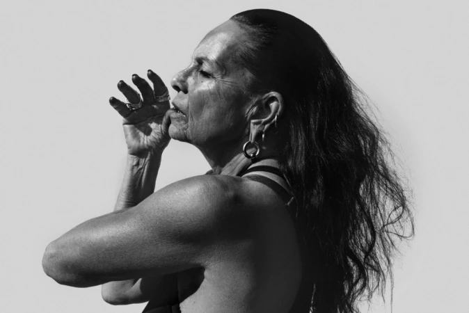 Monochrome profile portrait of an older woman with long dark hair, wearing rings, with hands raised.