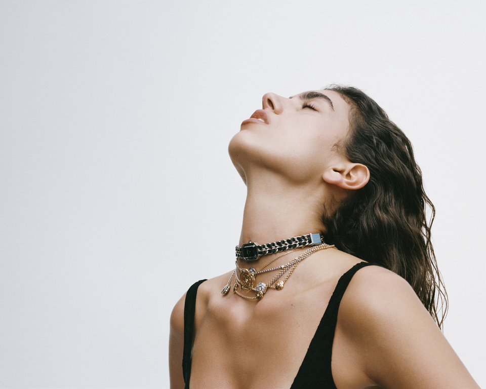 Portrait of a young woman with brown hair looking up and wearing decorative necklaces in a minimalist photo studio against a white backdrop.