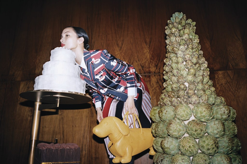 An Asian woman in a patterned blazer poses indoors next to a cake and artichoke tower, holding a yellow dog-shaped purse.