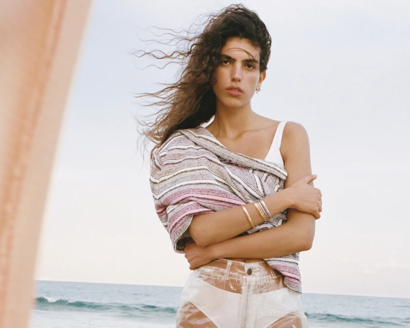 A woman stands on a beach with waves in the background, wearing a white top, sheer shorts, and a striped cover-up.