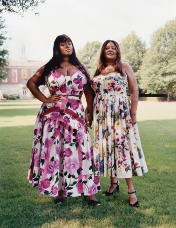 Two adult females stand in a grassy area, wearing floral dresses, under sunlight, with a building and trees in the background.